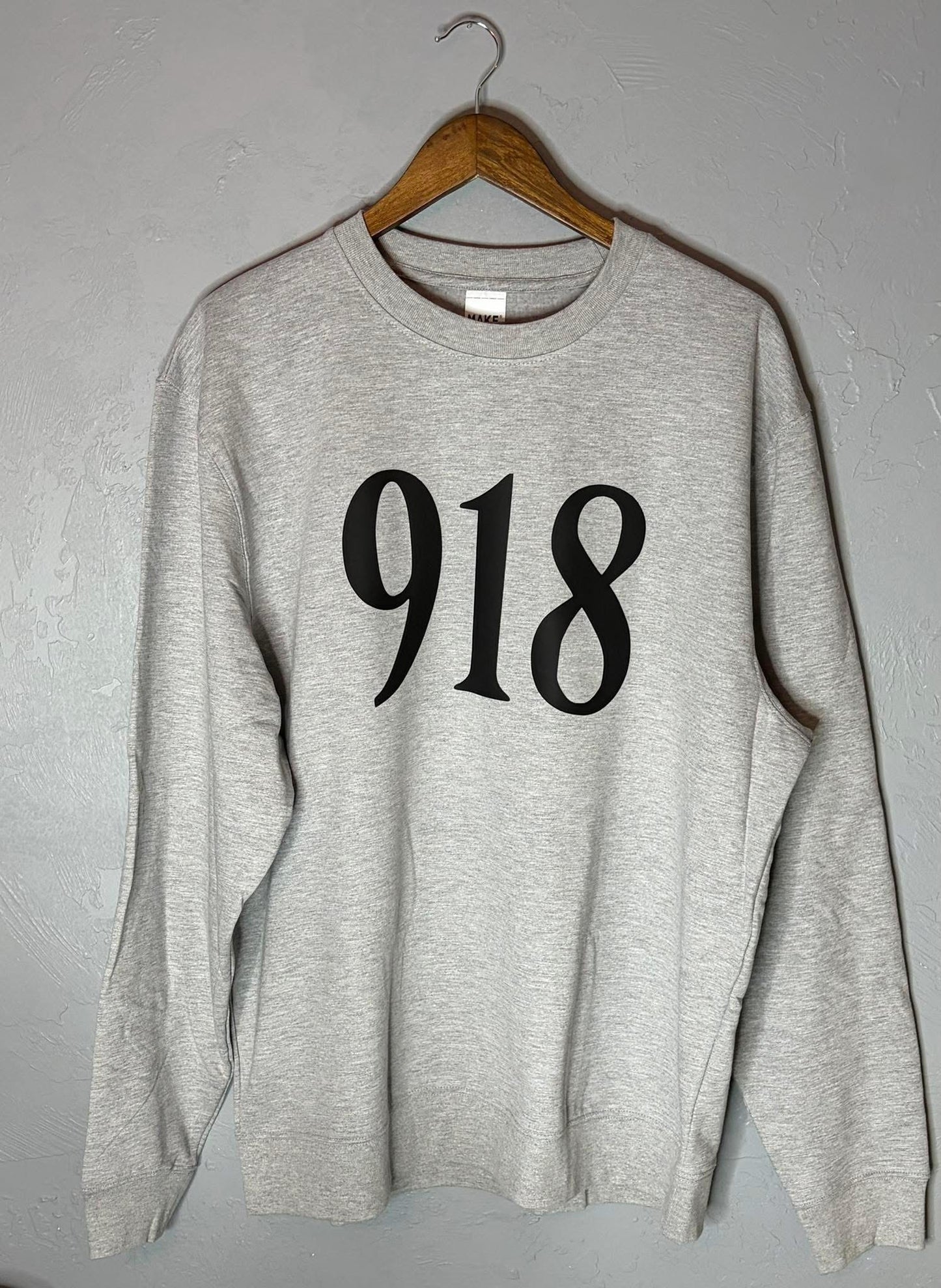 Gray sweatshirt with '918' printed on it hanging on a wooden hanger against a gray wall.