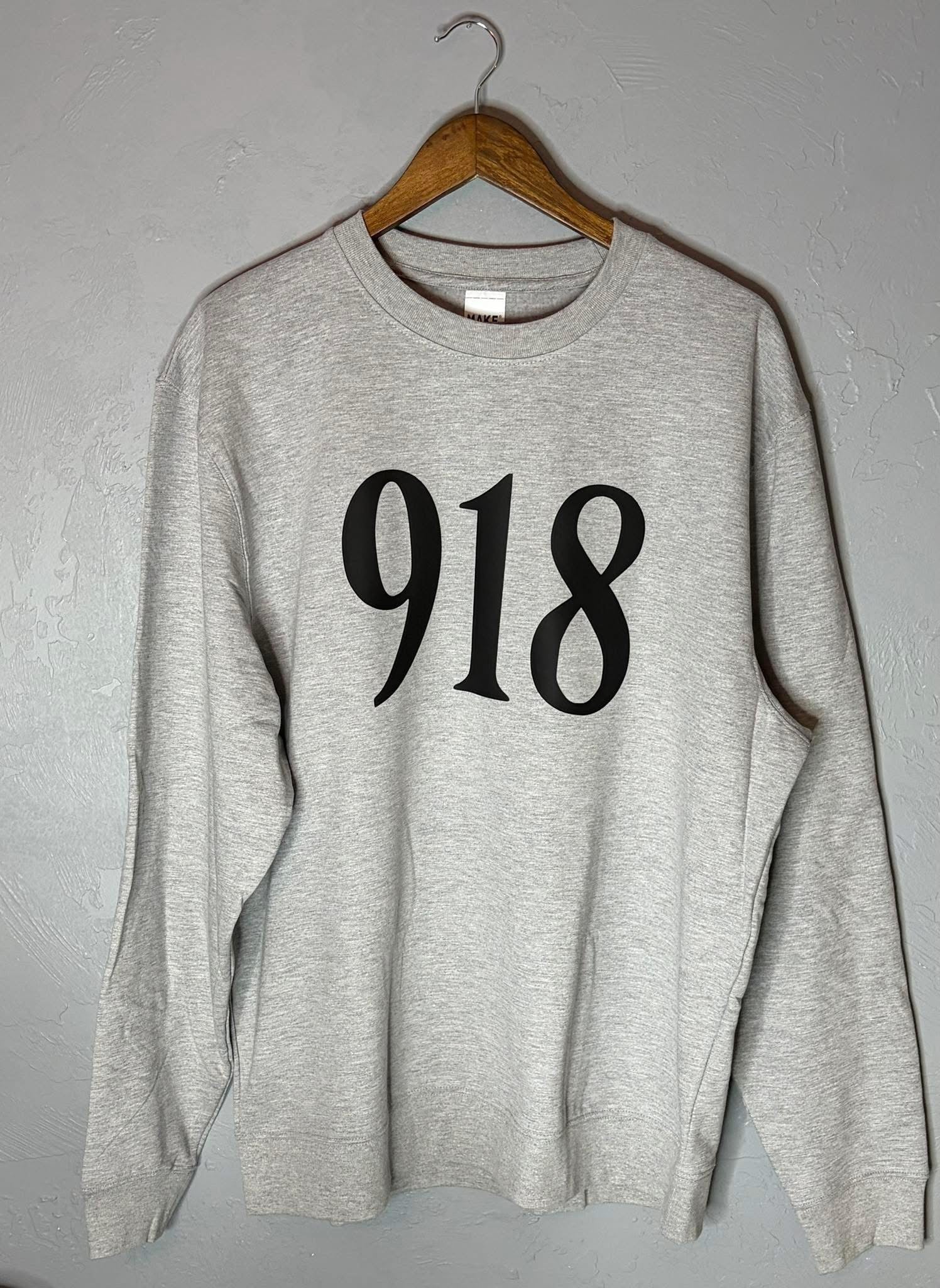 Gray sweatshirt with '918' printed on it hanging on a wooden hanger against a gray wall.